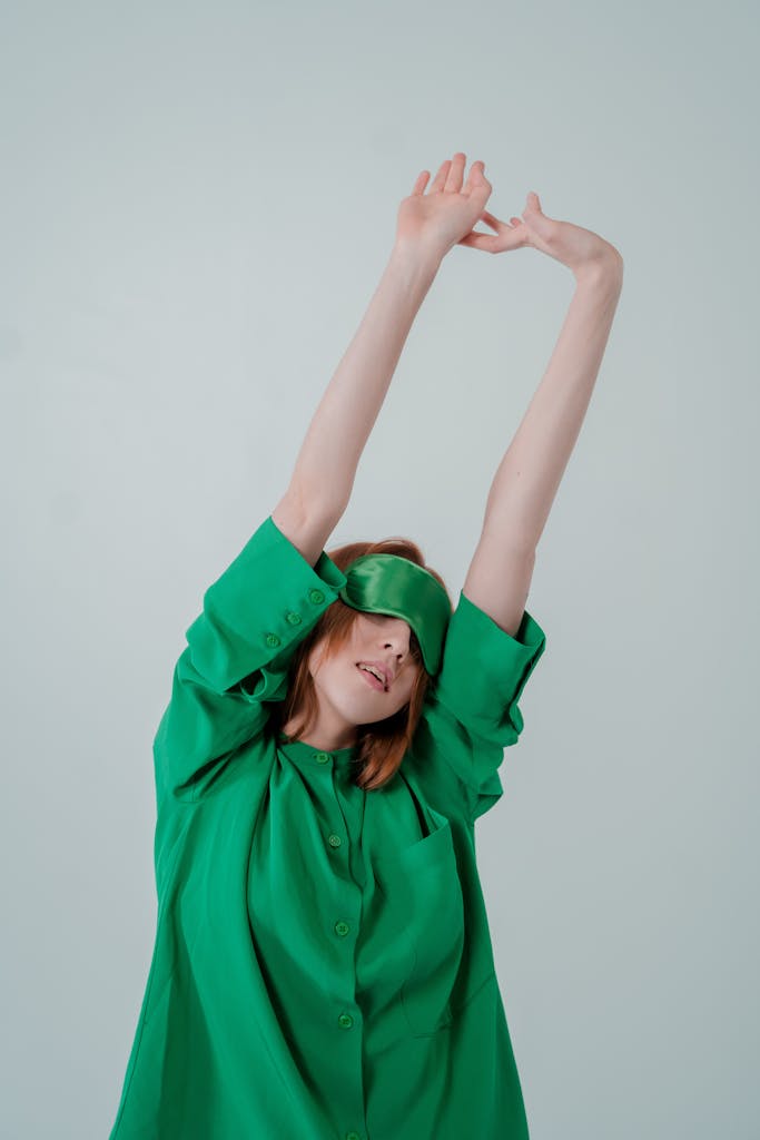 A woman in green sleepwear stretches with a sleep mask on a light background.